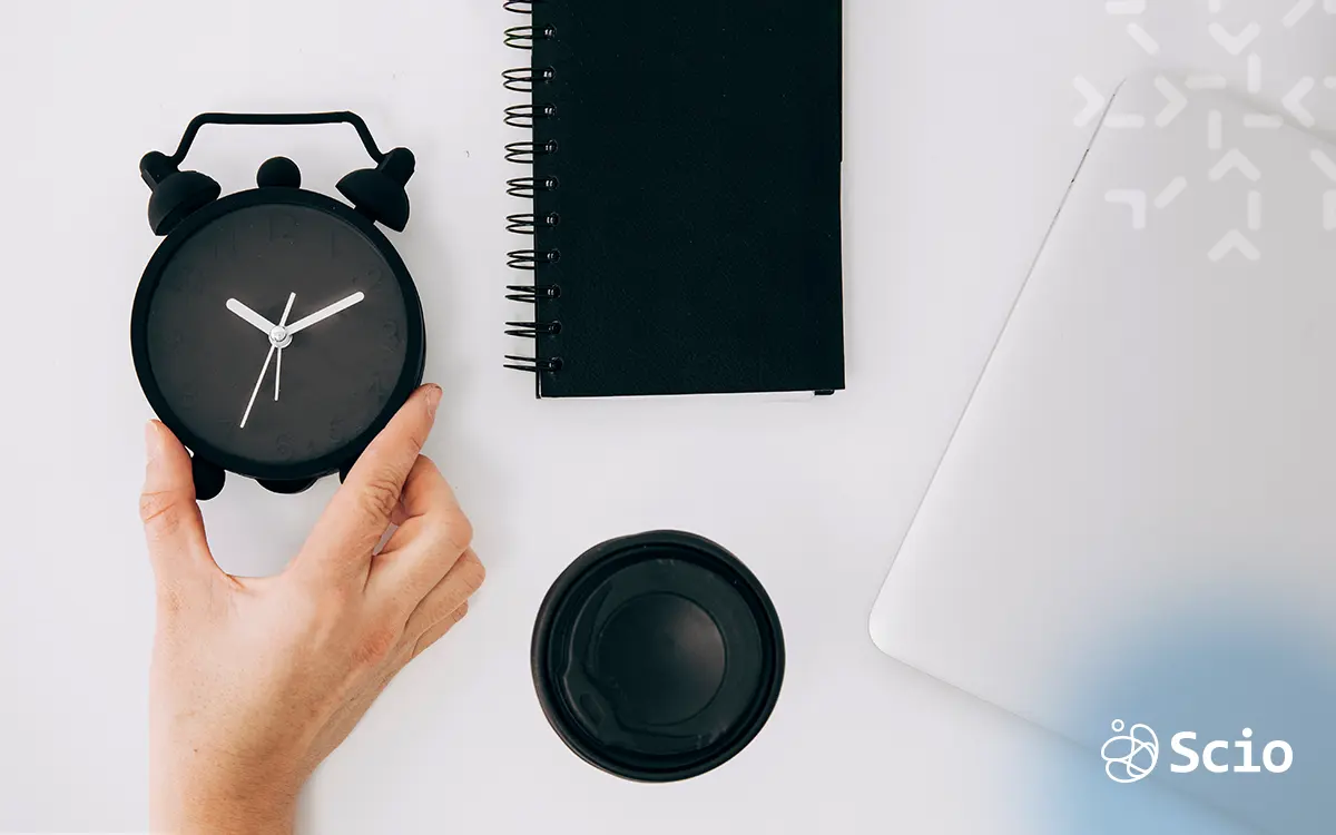 Visualizing Flexibility in Tech Work: Autonomy & Time Balance Top view of a person holding a black clock next to a blank notebook and laptop—symbolizing hybrid work, time autonomy, and modern work flexibility.