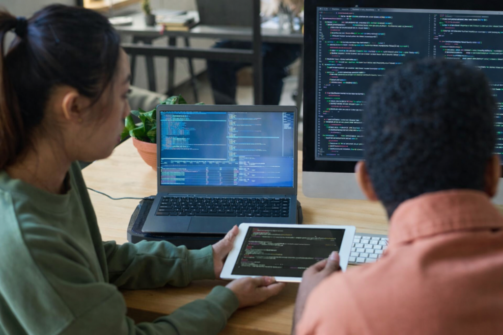 Software developers collaborating at a desk, reviewing code across multiple screens in a modern engineering workspace.